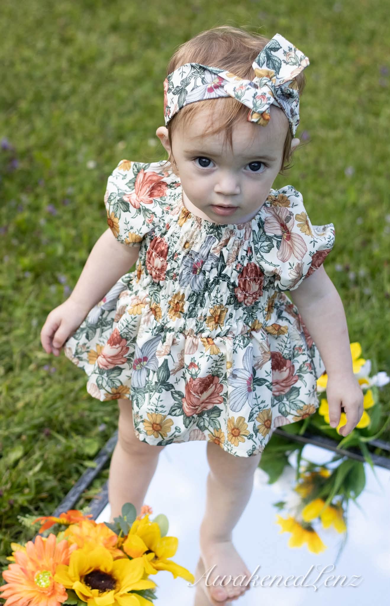 A toddler in a floral dress and headband standing beside sunflowers